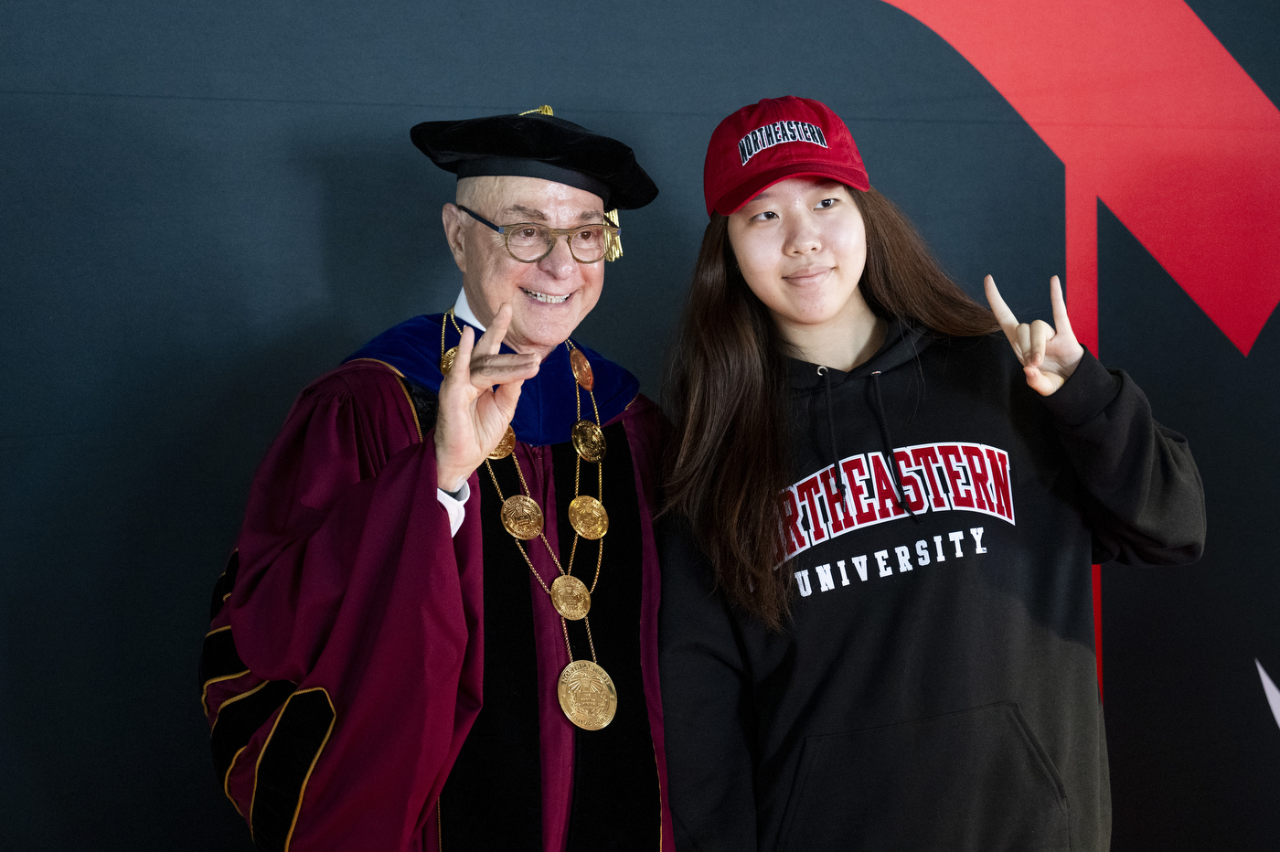 President Aoun in academic regalia poses with a student in a red cap.