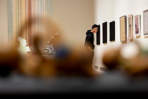 A student in a black hoodie studies a row of framed works on a gallery wall, seen past blurred objects in the foreground, with a tall multicolored hanging piece and small sculptures on a pedestal behind him.