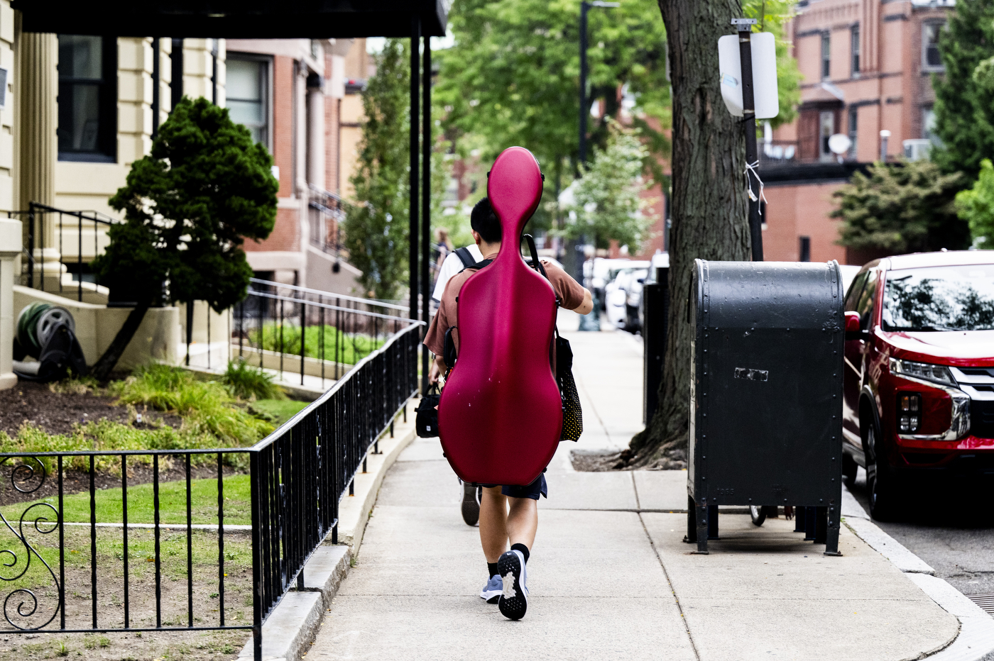 A student carries a cello case on their back while walking down a residential sidewalk.