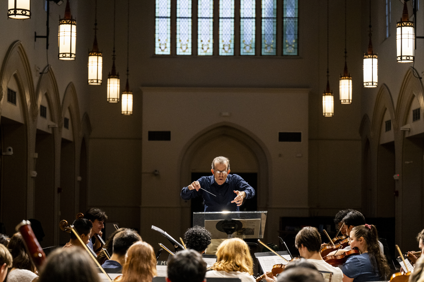 A conductor leads student musicians during rehearsal in a church-like hall with stained glass windows.
