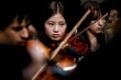 A student focuses while playing violin, with other musicians visible in soft focus around her.