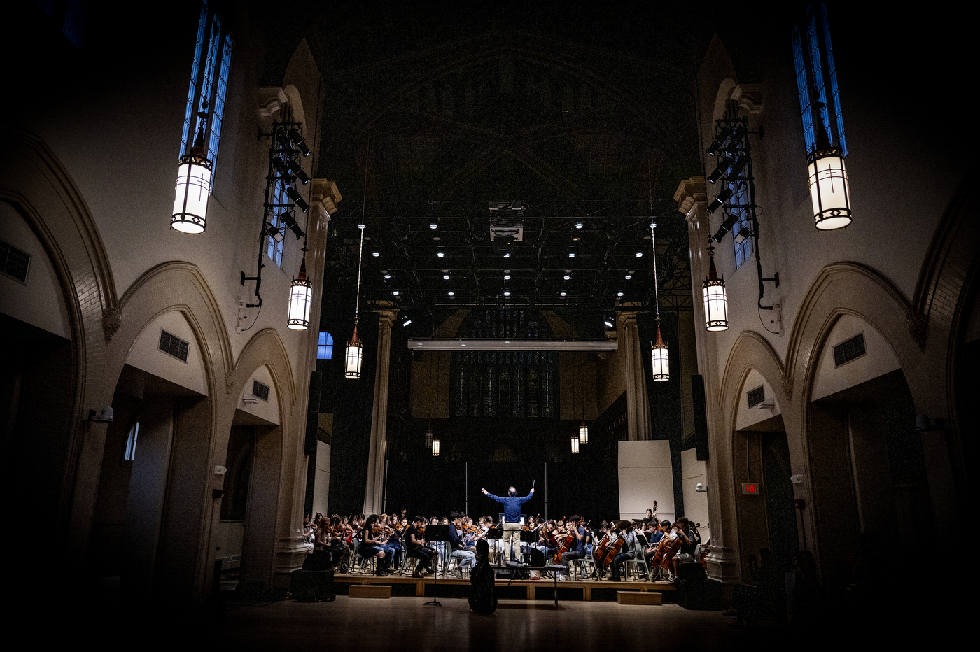 Wide view of the full orchestra rehearsing on a lit stage, framed by the dark, cathedral-like architecture of the hall.