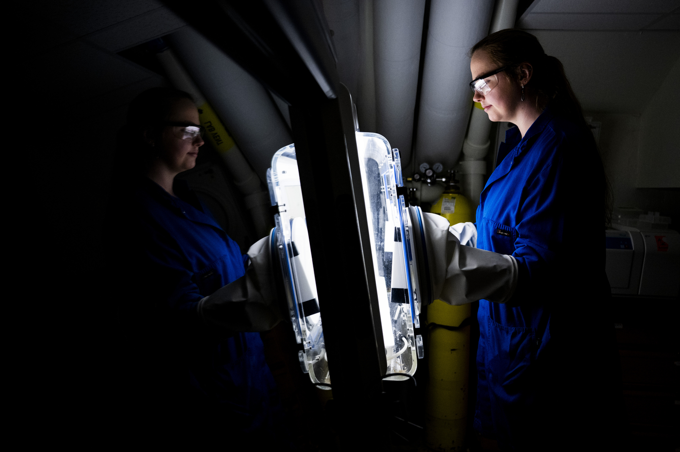 Two researchers in lab coats and safety goggles examine a large metallic apparatus in a darkened lab, illuminated by a handheld light.