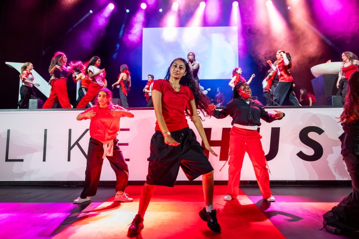 Performers in red dance on stage in front of 'Like Us' backdrop.