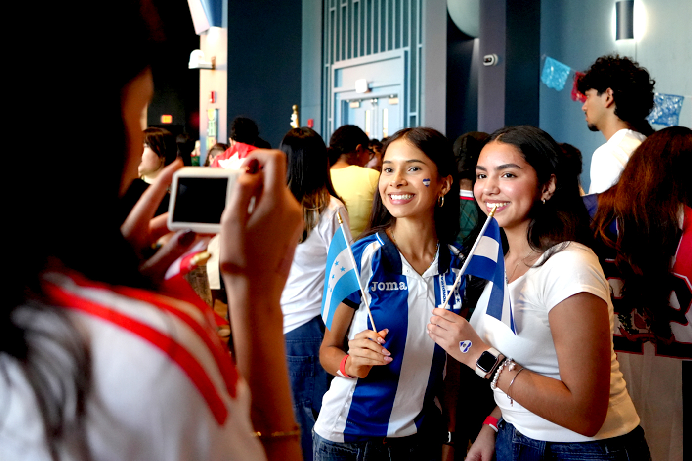Two students smile and pose with Honduran flags while another student photographs them, part of a Parade of Flags event celebrating cultural heritage.