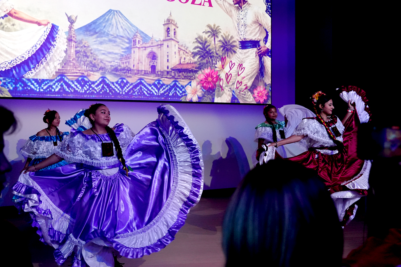 Students perform a traditional folk dance in colorful, full-skirted dresses on stage at the Parade of Flags event, with a projected image of a Central American landmark behind them.