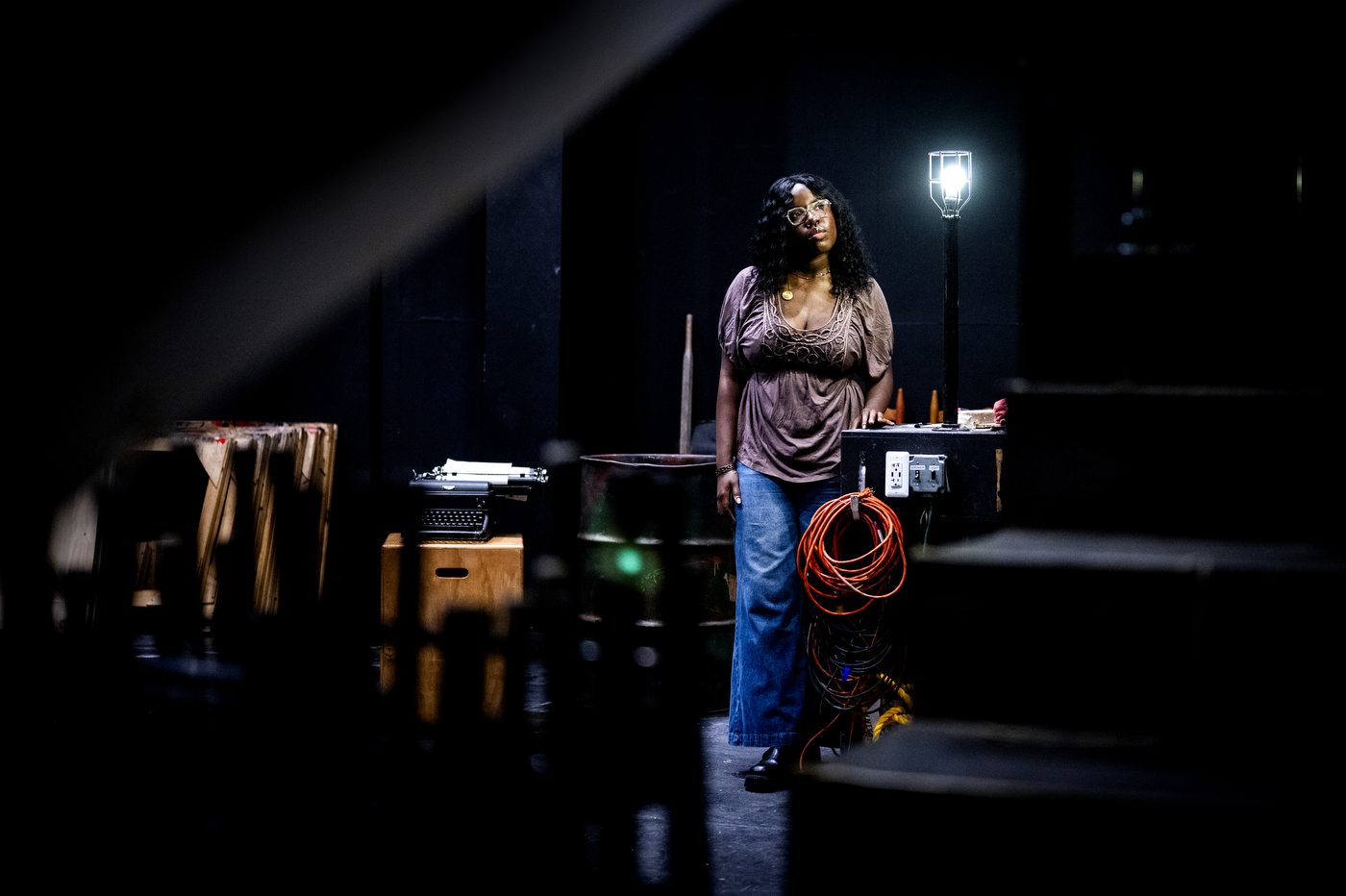 Rachel Oppong standing on a dark stage beside a light bulb, gazing upward, with a typewriter and a coiled orange extension