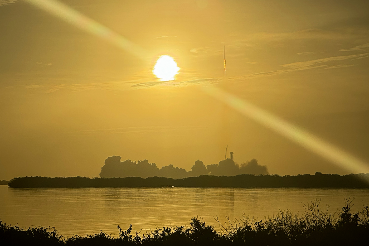 A rocket standing on its launch pad near the horizon at sunrise, seen across water with a bright sun overhead.