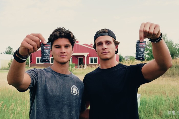 Northeastern alumni Andrew Leisengang and Alex Jimenez hold up sachets of their energy honey in a grassy field in front of a red barn-style building.