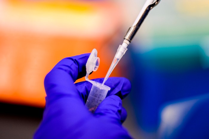 Gloved hand in blue nitrile holding a pipette over lab equipment, with a blurred bioinformatics-themed display in the background.