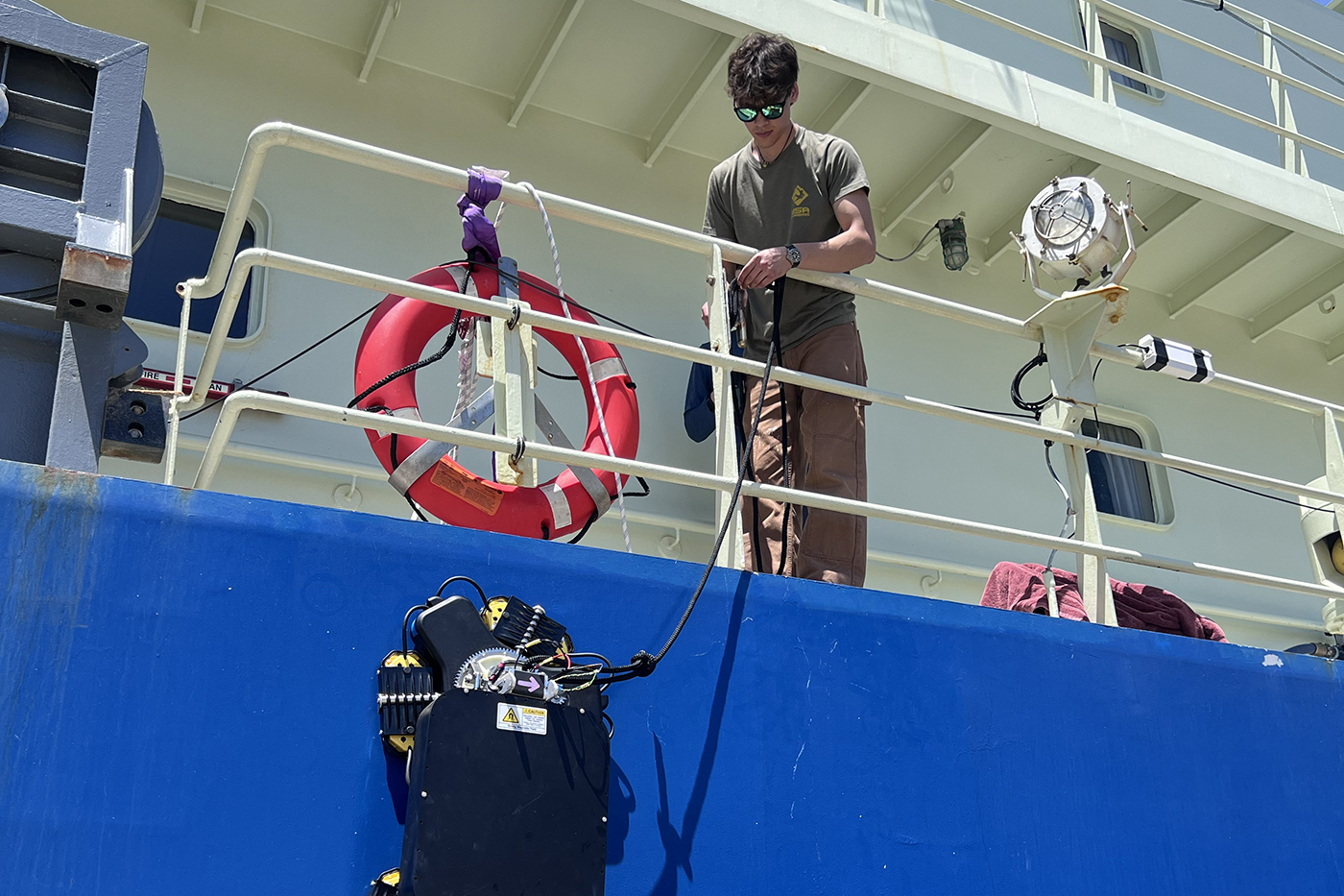 A person on the deck of a large ship lowers a robotic device over the side by a cable, beside an orange life ring on the
