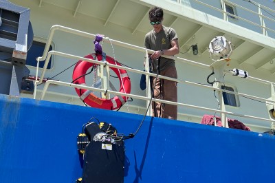 A person on the deck of a large ship lowers a robotic device over the side by a cable, beside an orange life ring on the railing.