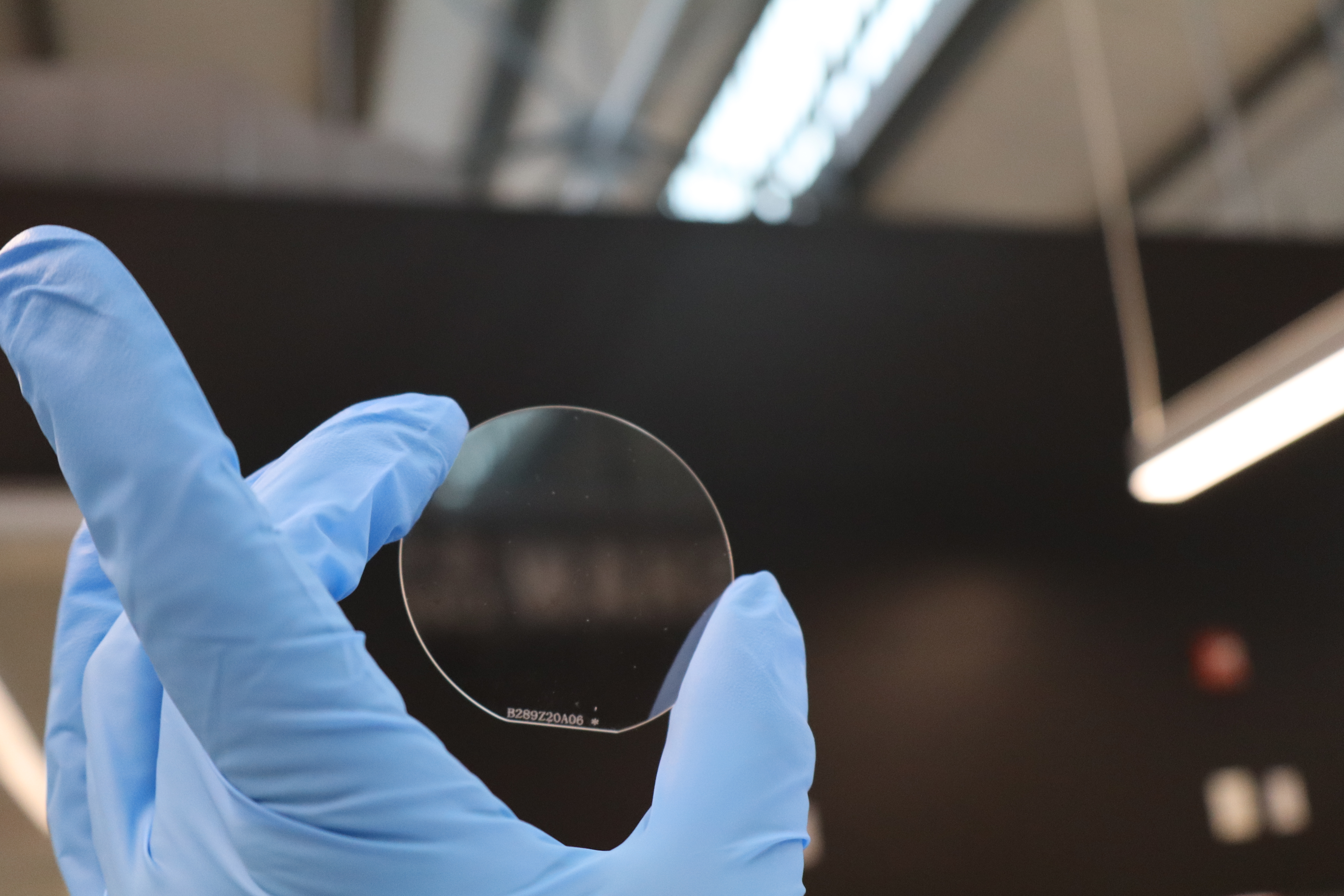 A gloved hand holds a thin, transparent circular wafer up for inspection in a lab.