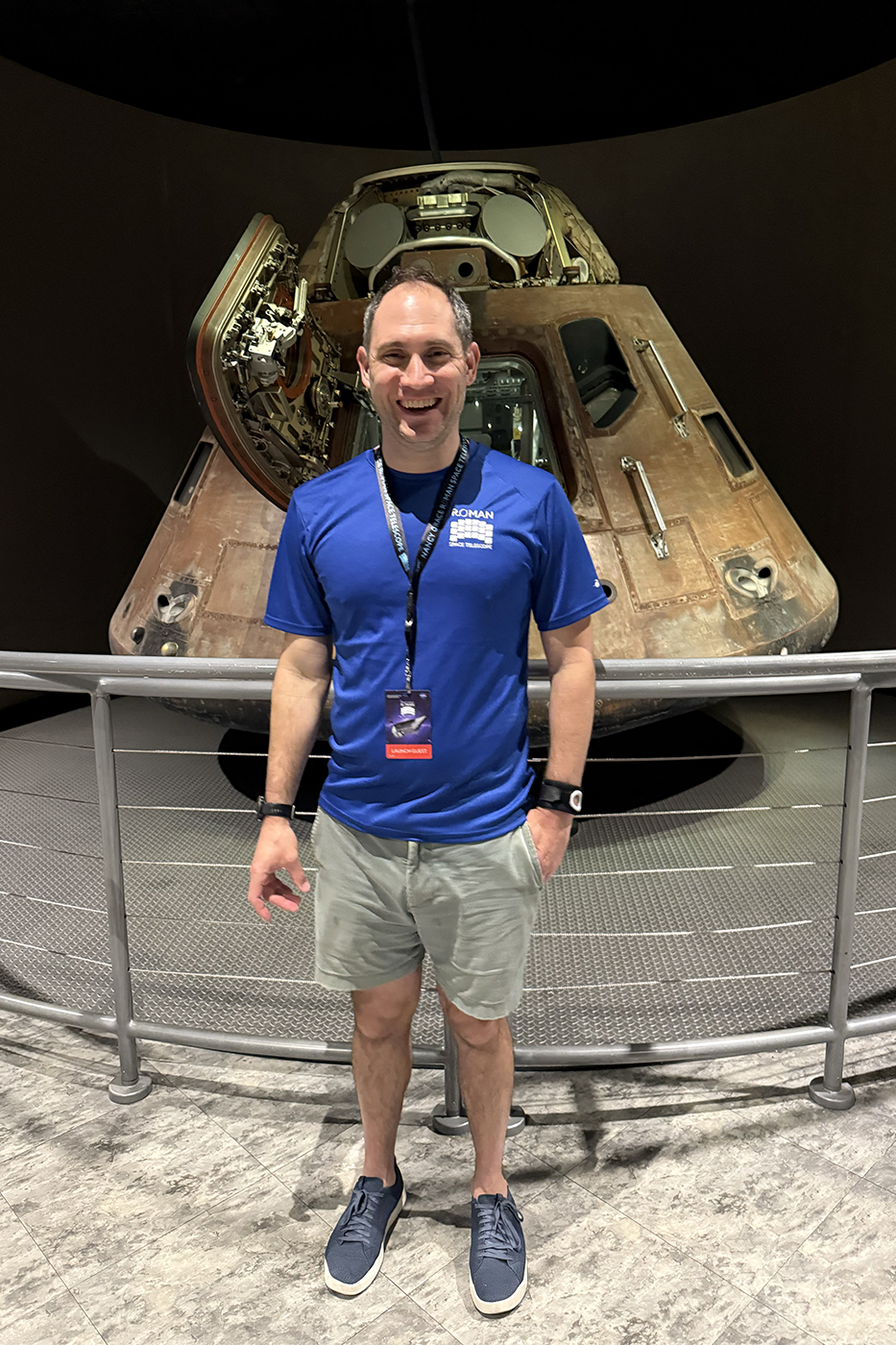 A man in a blue T-shirt and shorts smiling in front of a retired space capsule on display.