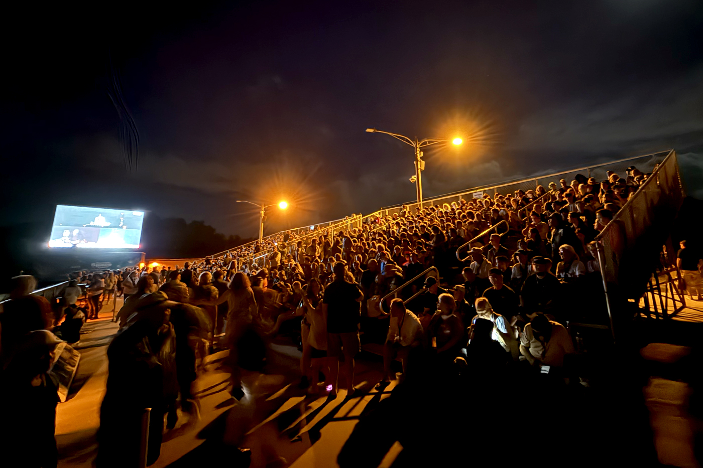 A large crowd gathered along a waterfront at night to watch a launch, with a screen and lights visible in the distance.