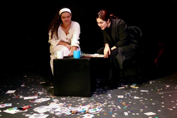 Two young women sit at a small black table on a stage scattered with confetti and photographs, one leaning over to look at something the other is holding.