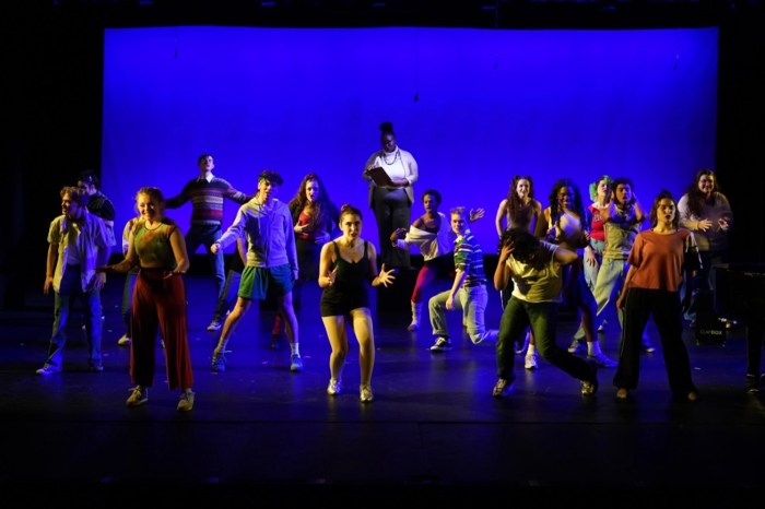 A large cast of student performers dances in a crouched, energetic pose on a stage lit in blue, with a woman in a black tank top and shorts at center and a performer holding a clipboard standing behind them.