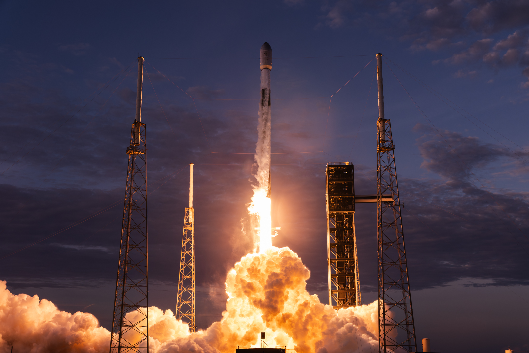 A rocket lifts off from a launch pad at dusk, engulfed in exhaust flame and smoke.
