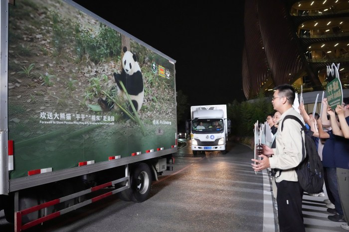 At night, people lining the roadside hold up signs and phones as a truck printed with a panda image and a farewell message drives past, with a second truck following behind.