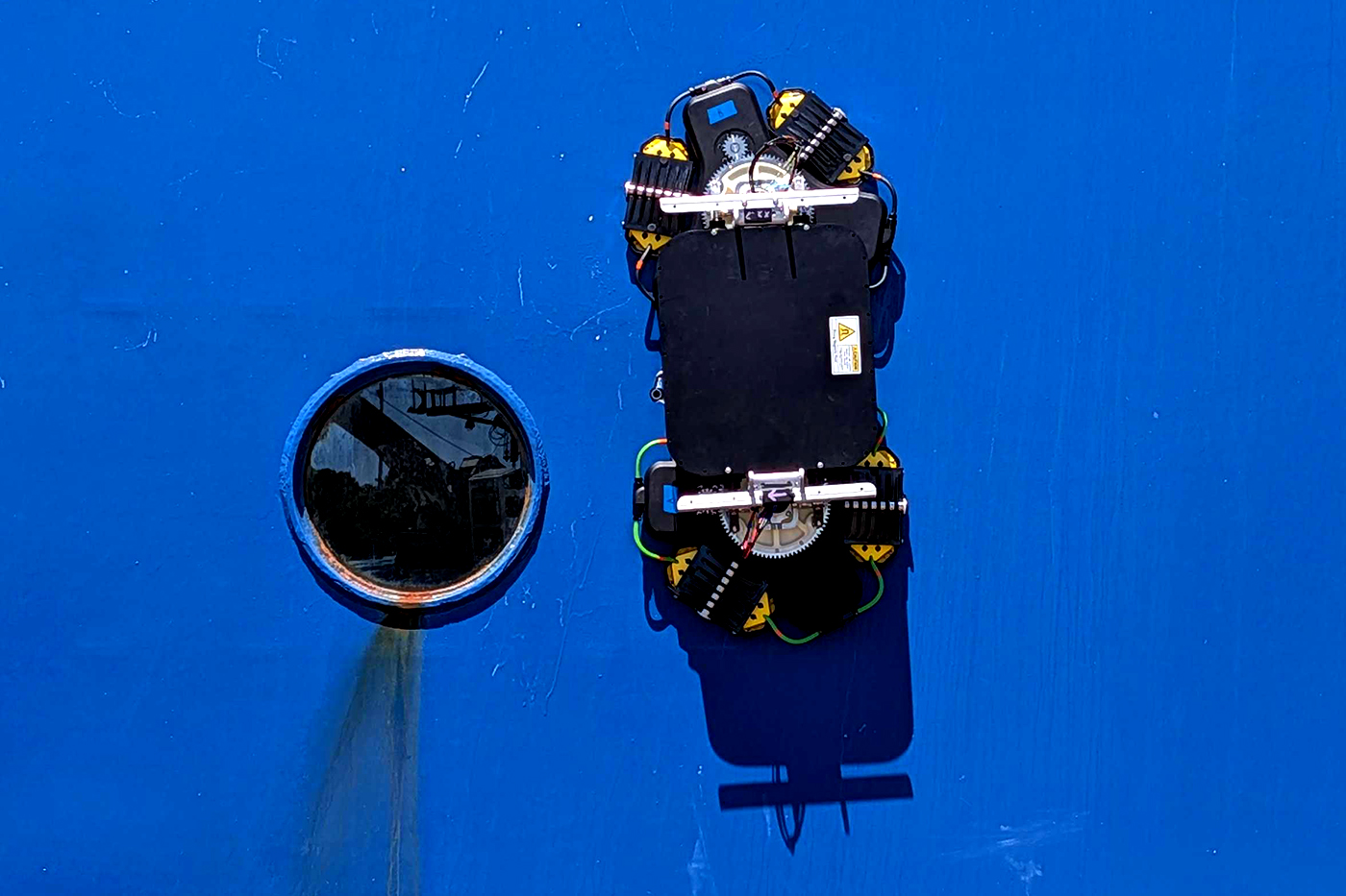 A robotic device with wheeled tracks attached to the blue hull of a ship, next to a porthole.