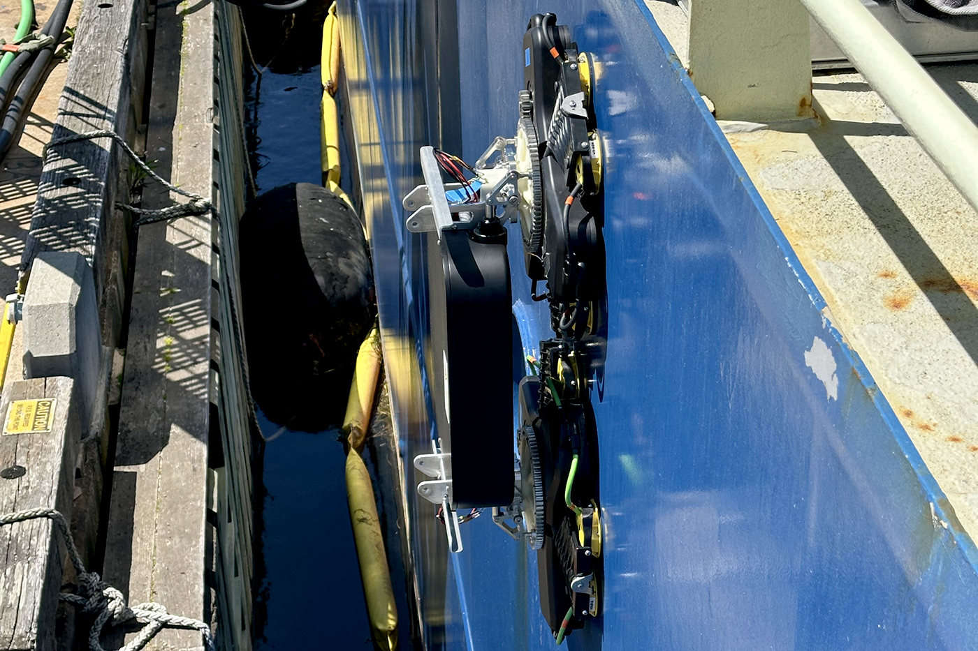 A robotic device on the side of a ship's blue hull, seen from above, in the narrow gap between the ship and a wooden dock lined with fenders.