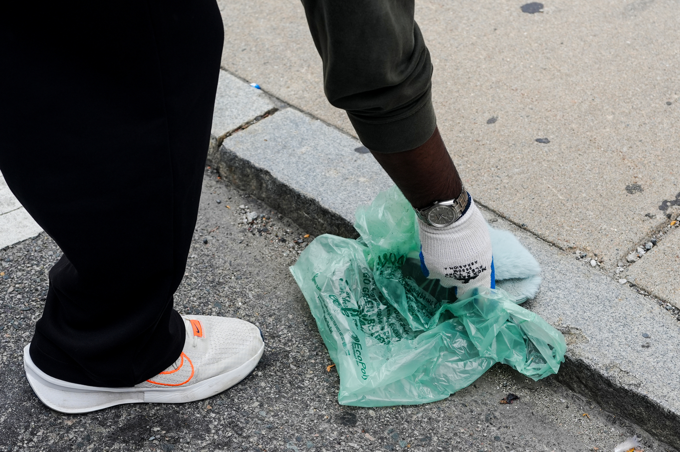 A person bends down to pick up trash from a sidewalk using a plastic bag.