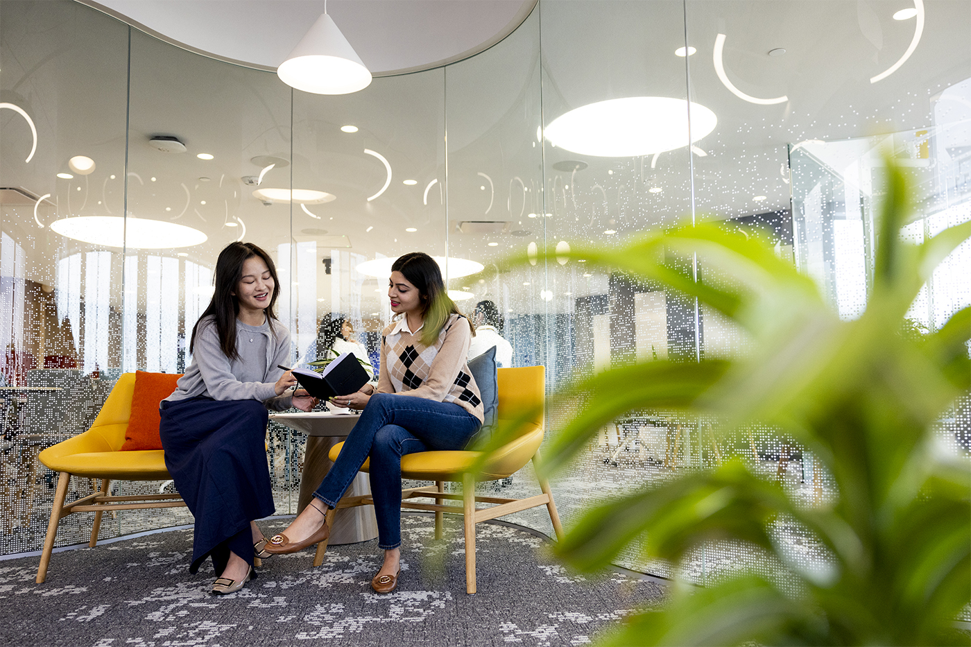 Two students talk at a small table in a glass-walled study room at Northeastern’s Silicon Valley campus, one holding an open notebook.