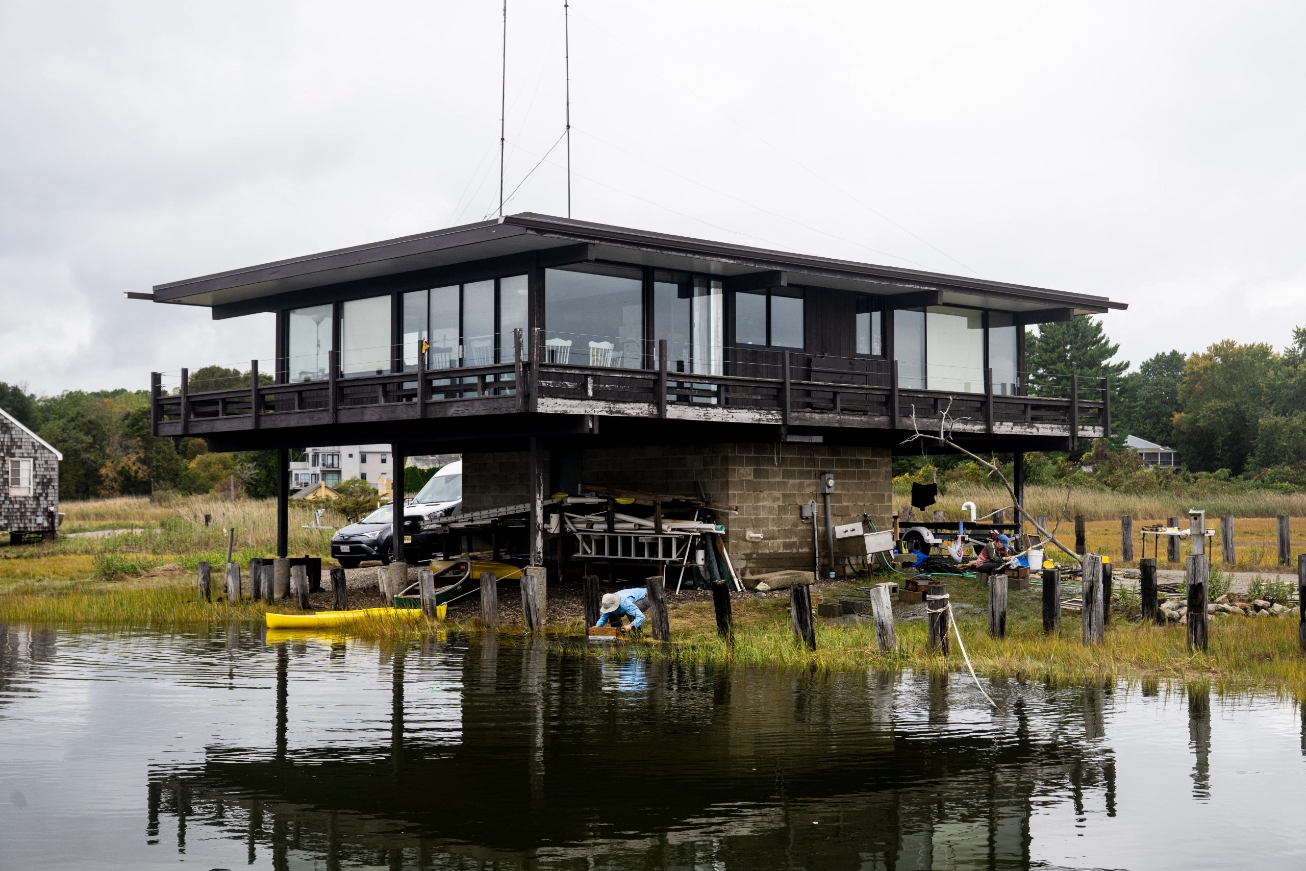 A far away shot of a researcher crouching to pull up clams, underneath a large building on a foundation above the water.