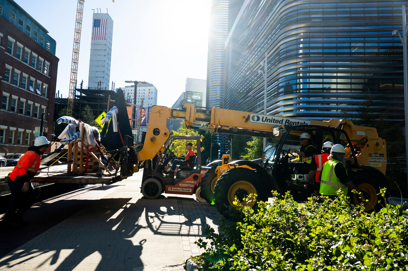 Crew members in hard hats and safety vests use a telehandler and forklift to unload a sculpture wrapped in padding and straps from a flatbed truck.