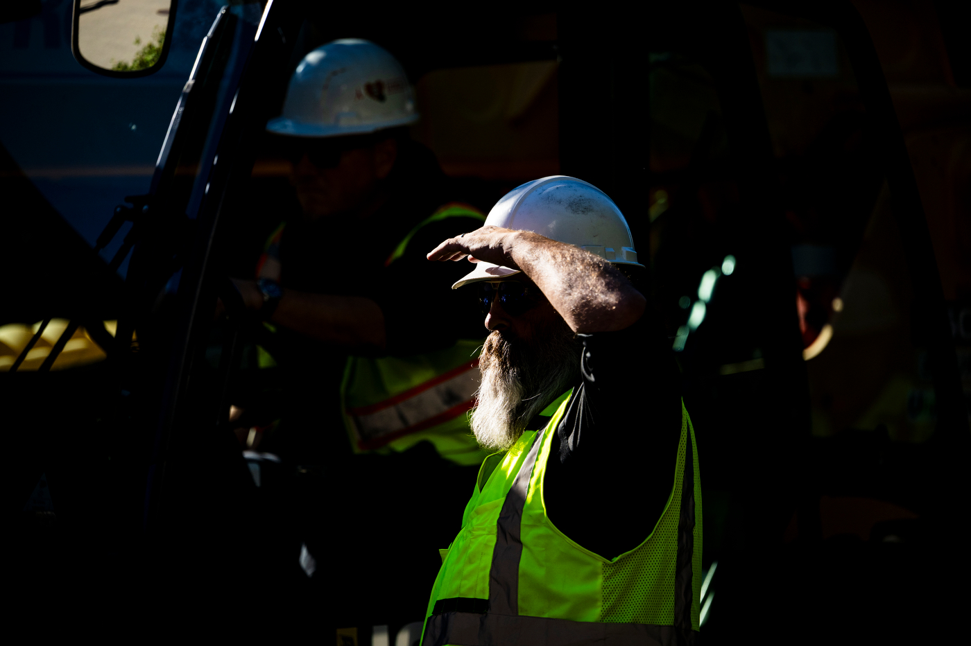 A bearded worker in a white hard hat and yellow safety vest shields his eyes with his hand while standing in deep shadow.