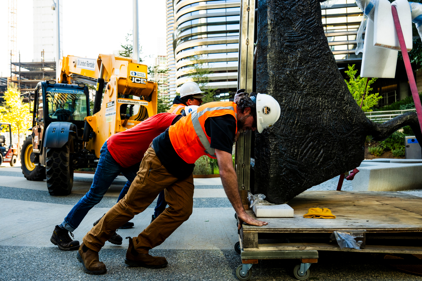 Two workers in hard hats lean into a wheeled wooden platform, pushing a large, dark, textured section of a sculpture across a plaza.