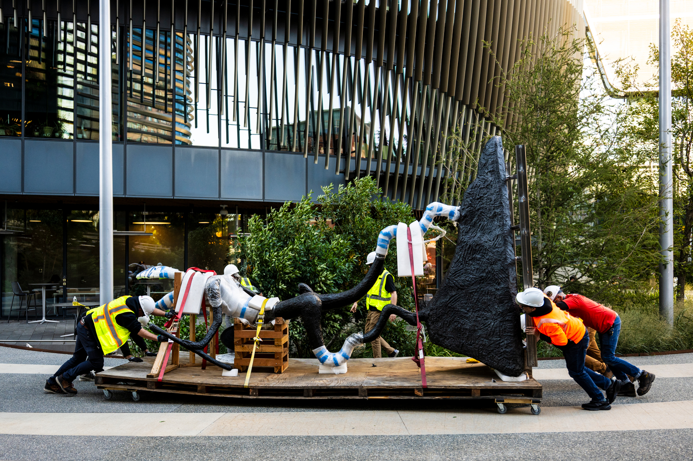 A crew pushes a long wheeled platform carrying a dark sculpture laid on its side, its thin limbs wrapped in padding and straps.