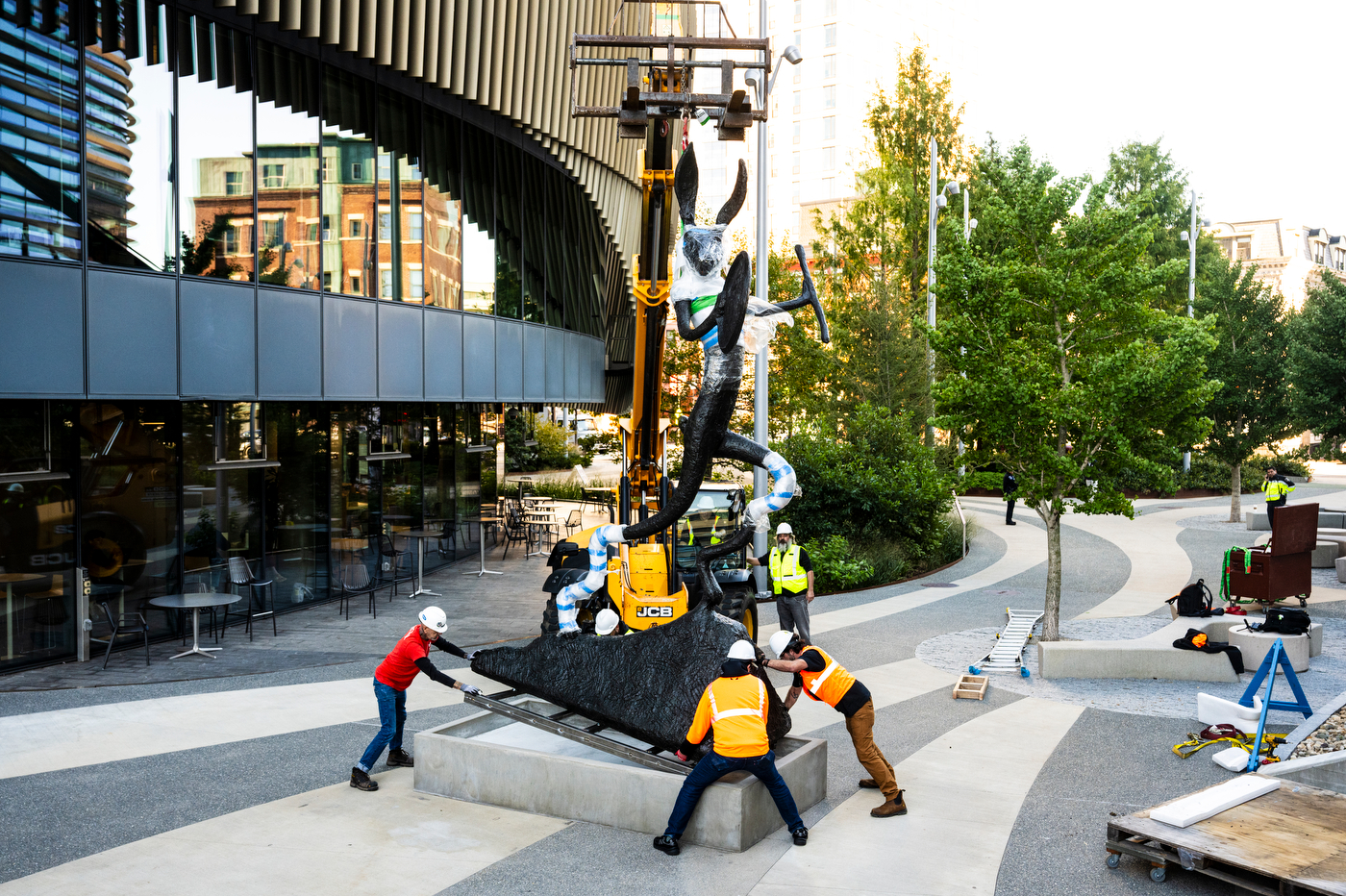 Crew members guide the base of the Large Left-Handed Drummer onto a concrete pedestal as a telehandler holds the upright, partly wrapped hare sculpture in place on a campus plaza.