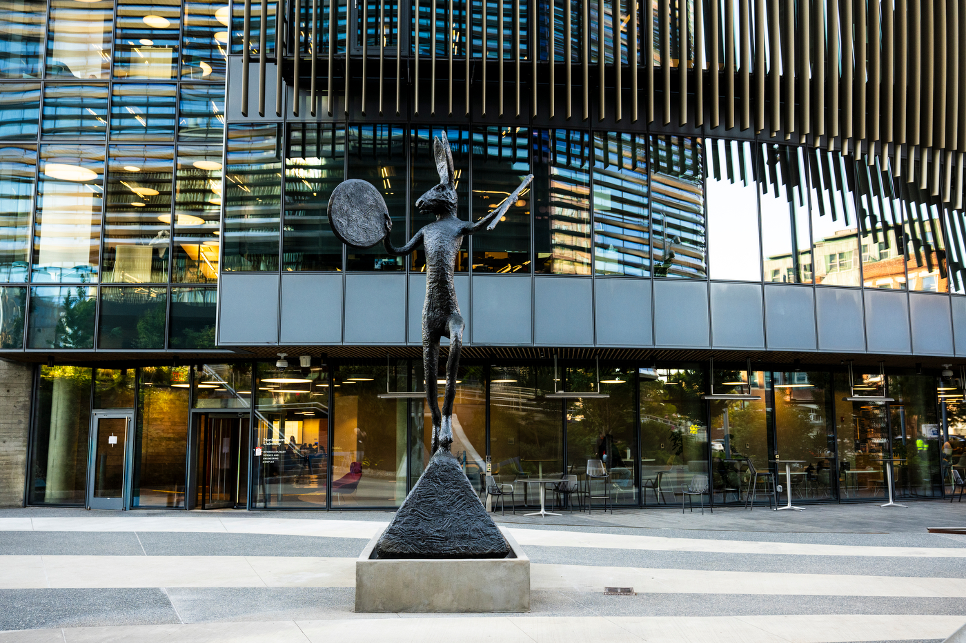 The Large Left-Handed Drummer, a tall bronze hare raising a drum and drumstick on top of a pyramid-shaped base, installed in front of the glass facade of Northeastern's Interdisciplinary Science and Engineering Complex.