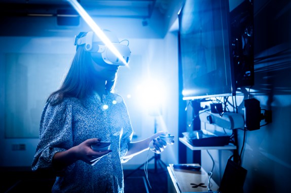 A student wearing a virtual reality headset holds controllers while using a VR system in Northeastern University’s Immersive Media Lab.