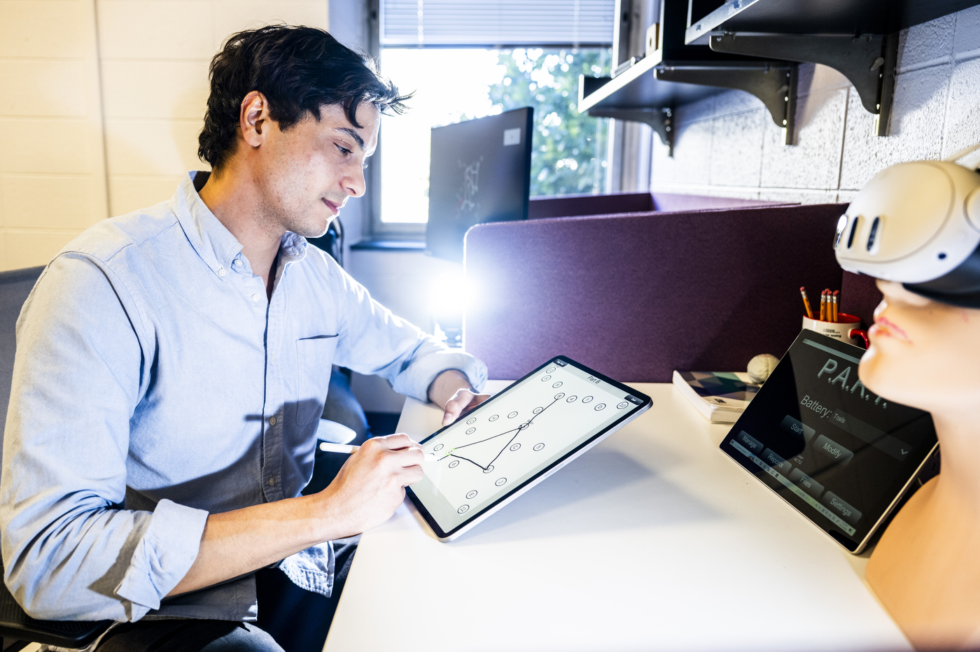 Quentin Coppola looking at a tablet while seated next to a participant wearing a virtual reality headset in an office. 