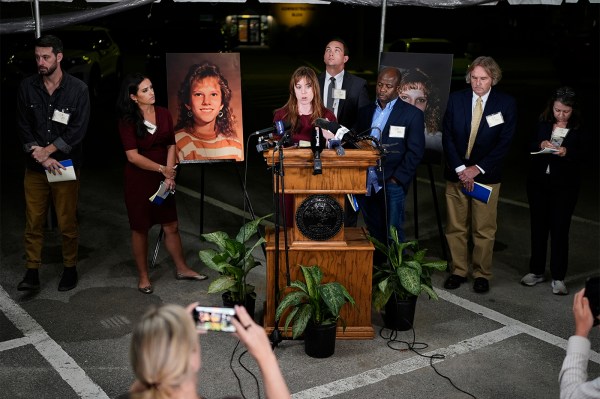 State officials and members of the media gather around a podium in a dimly lit room.