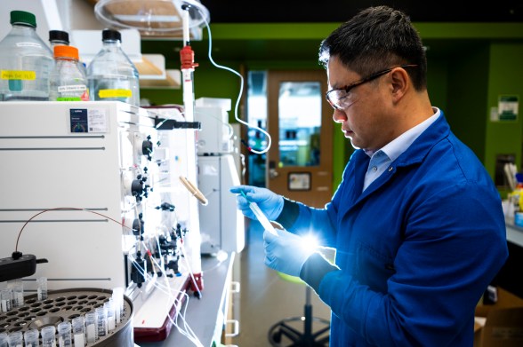 A researcher in a blue lab coat, safety glasses and gloves examines a small glowing tube beside a lab instrument fitted with reagent bottles, tubing and a tray of sample vials.