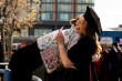 A laughing graduate in a black cap and gown, holding a card, hugs someone wearing an embroidered patterned stole outdoors on a sunny day as other graduates gather in the background.