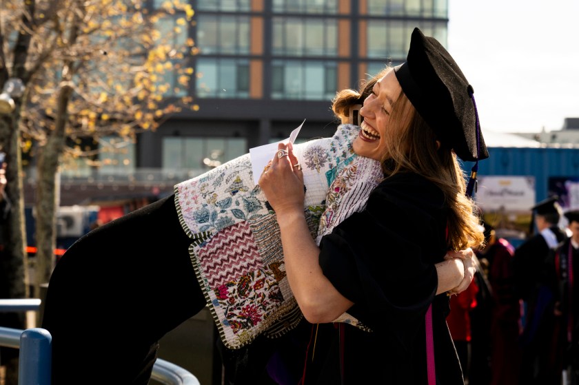 A laughing graduate in a black cap and gown, holding a card, hugs someone wearing an embroidered patterned stole outdoors on a sunny day as other graduates gather in the background.