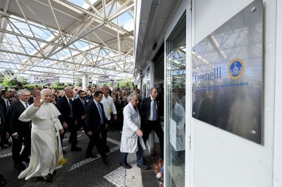 Pope Leo XIV waves while arriving at RomeâÂÂs Gemelli University Hospital, accompanied by officials and medical staff.