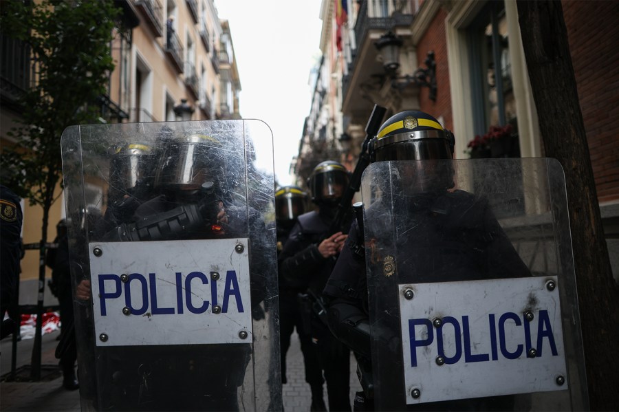 Police in riot gear caay shields that say "Policia" in a European city.