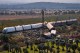 Elevated view of a derailed white high-speed train beside red-and-white passenger cars that lie jackknifed and overturned on an embankment strewn with debris, with fire trucks and rescue crews gathered in an orchard below.