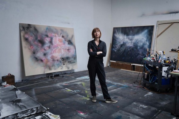 Yulia Pinkusevich standing with arms crossed in her paint-spattered studio, between a large pink and gray abstract canvas on the left and a dark, smoky painting on the right, with a cart of paints and supplies nearby.