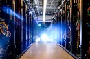 A person standing at a cart at the end of a narrow aisle between rows of server racks filled with colored cables, lit by a bright blue-white glow.
