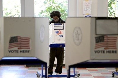 A person with a dark skin tone and glasses is hunched over a voting booth casting a ballot.