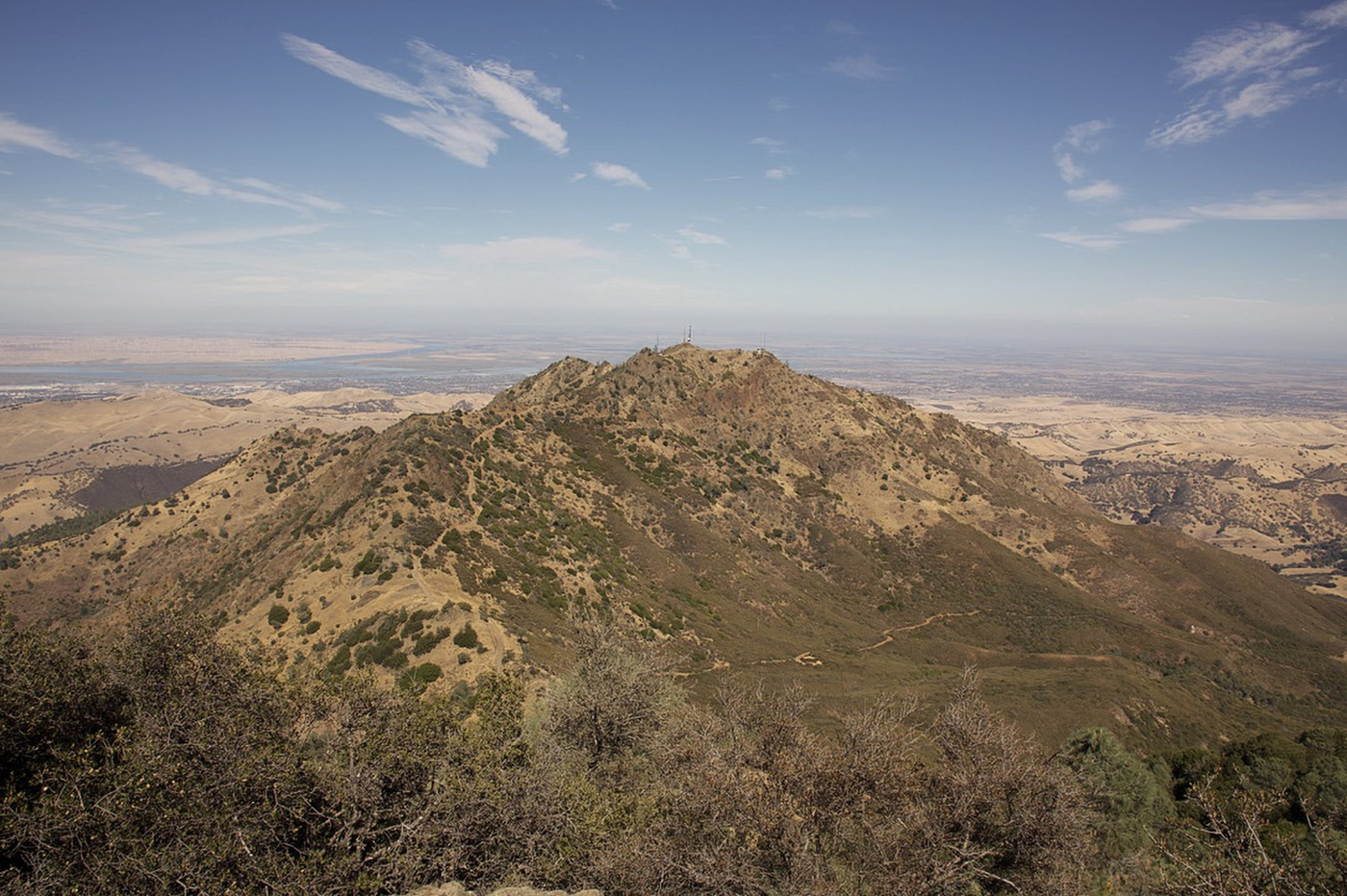 A rocky peak with dry, golden slopes and patches of green scrub, a wide valley stretching to the horizon behind it.