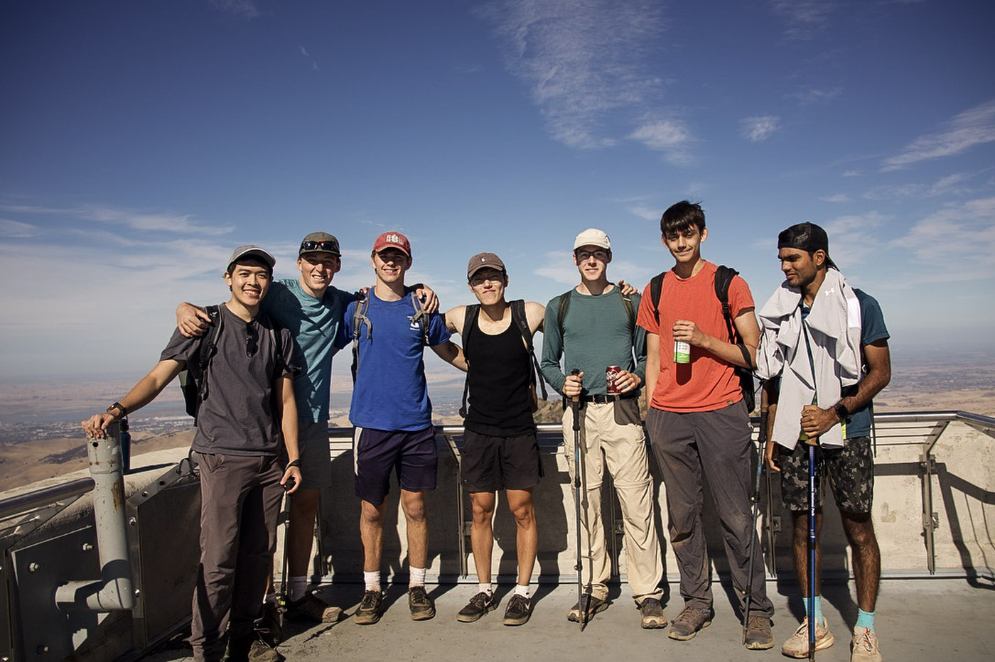 Seven hikers with backpacks posing arm in arm at a railed summit lookout, with a wide valley and a hazy horizon behind them.
