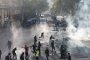 Protesters run through clouds of tear gas as police and armored vehicles stand in the background on a tree-lined street in France.