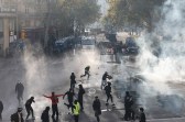 Protesters run through clouds of tear gas as police and armored vehicles stand in the background on a tree-lined street in France.